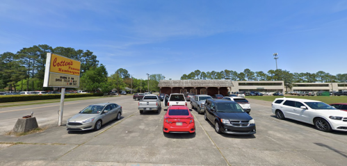 A parking lot with various cars, a sign for "Cotton's" in the foreground, and a building in the background under a clear blue sky.