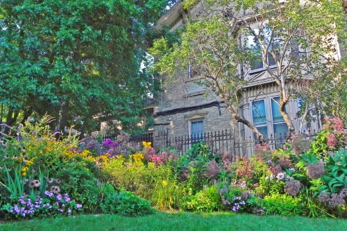 A charming stone house surrounded by vibrant flowers and lush greenery in a sunny garden setting.
