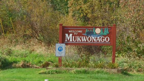 A welcome sign for Mukwonago, surrounded by greenery and trees, with a blue circular emblem nearby.