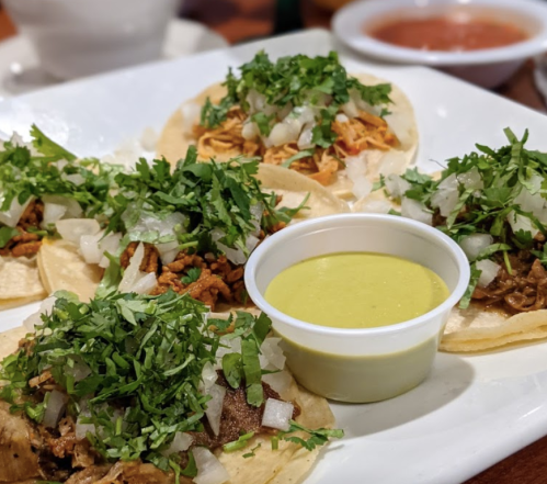 A plate of tacos topped with cilantro and onions, served with a small cup of green sauce and a side of salsa.