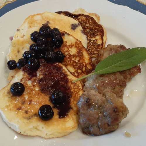 Plate of pancakes topped with blueberries and syrup, alongside a serving of sausage and a fresh sage leaf.
