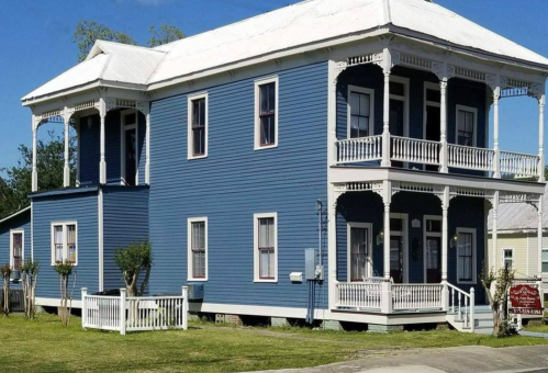 A large blue two-story house with white trim, featuring porches and a well-maintained yard.