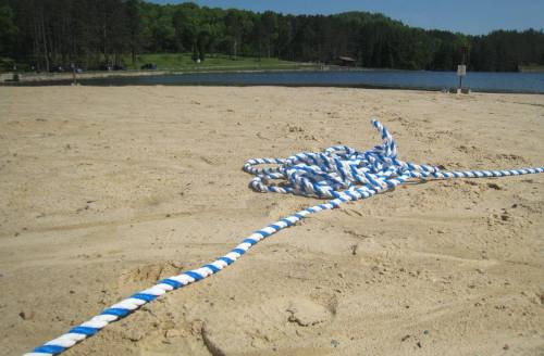 A coiled blue and white rope lies on sandy beach near a calm lake surrounded by green trees.
