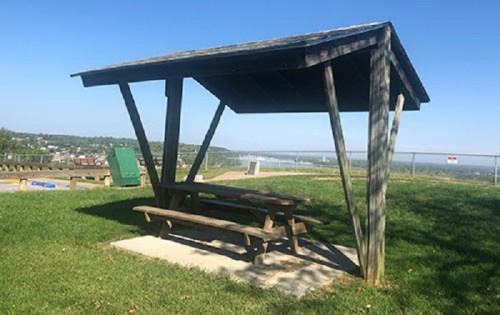 A wooden picnic shelter with a table, set on a grassy area overlooking a scenic view. Clear blue sky above.