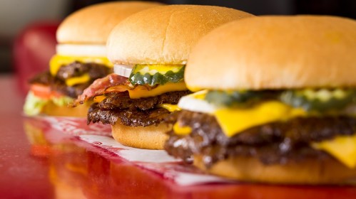 Three delicious cheeseburgers with lettuce, tomato, pickles, and bacon on a red table.
