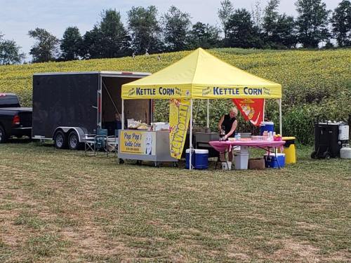 A kettle corn stand with a yellow tent, surrounded by fields, and a vendor preparing snacks.