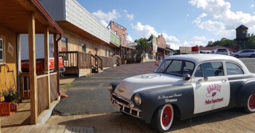 A vintage police car parked in front of a rustic storefront with blue skies and clouds in the background.