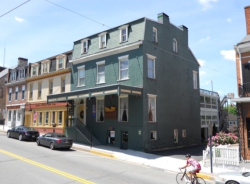 A green three-story building next to a yellow building, with a cyclist passing by on a sunny day.