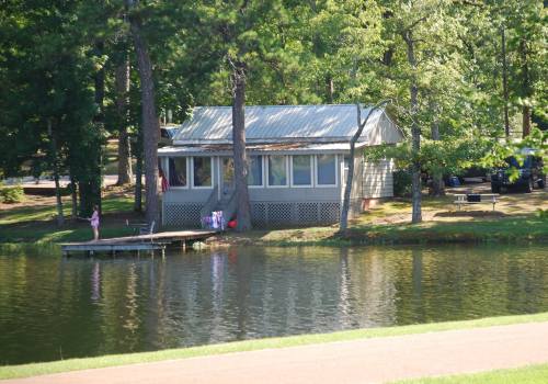 A small house by a calm lake, surrounded by trees, with a dock and a person standing nearby.