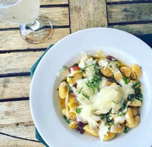 A bowl of gnocchi topped with cheese and herbs, served with a glass of drink on a wooden table.