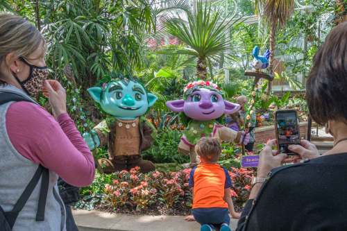 Two colorful, cartoonish characters with floral crowns stand in a lush garden, while visitors take photos nearby.