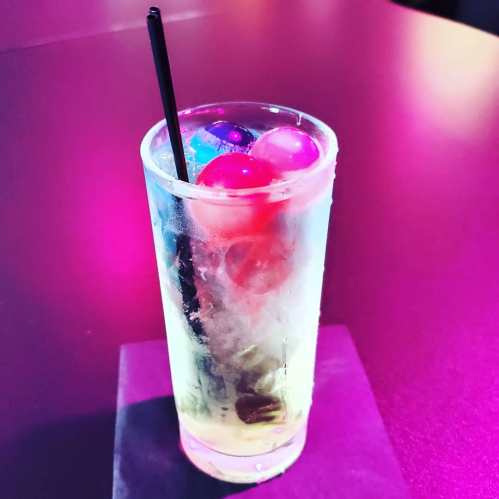 A clear cocktail glass filled with ice and colorful round fruit garnishes, set against a vibrant purple background.