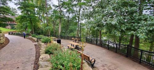 A scenic pathway lined with benches and greenery, leading to a river surrounded by trees.