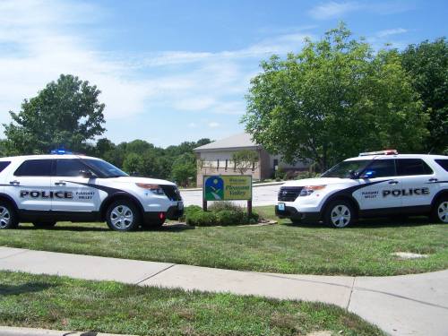 Two police SUVs with lights on parked near a sign that reads "Welcome to Pleasant Valley" on a sunny day.