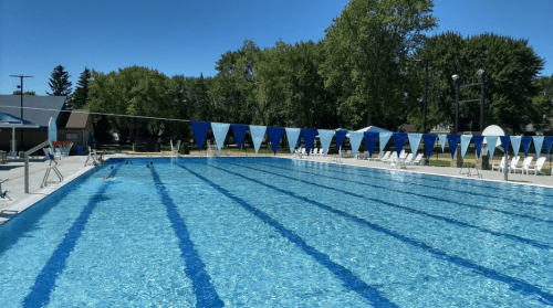 A sunny outdoor swimming pool with clear blue water, lane markers, and trees in the background.