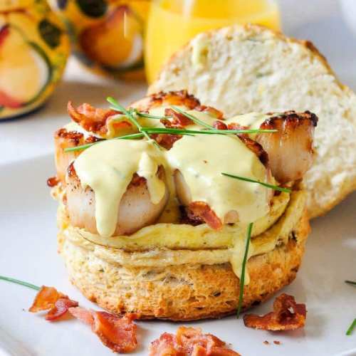 A toasted bun topped with scallops, bacon, and drizzled with creamy sauce, served on a white plate.