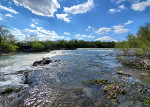 A scenic view of a river with a waterfall, surrounded by lush greenery and a bright blue sky with fluffy clouds.