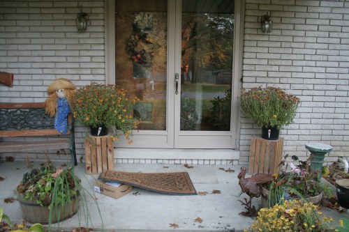 A cozy front porch with potted flowers, a scarecrow, a welcome mat, and a decorative rooster.