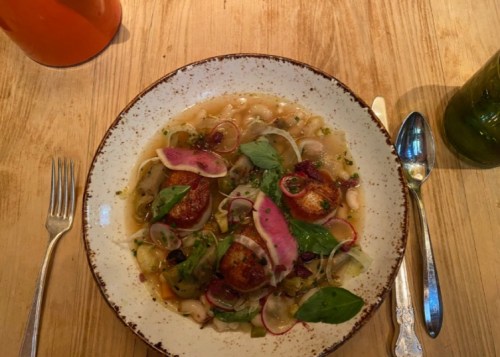 A plate of scallops with beans, greens, and radish, garnished with herbs, on a wooden table.