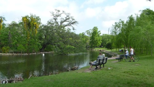 A serene park scene with a pond, benches, and people walking and biking along a path surrounded by lush greenery.