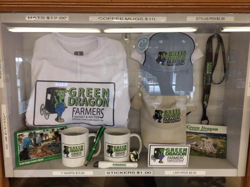 Display case featuring merchandise from Green Dragon Farmers Market, including shirts, hats, mugs, and pens.