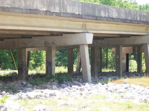 Underneath a concrete bridge, with support pillars and rocky ground, surrounded by greenery and graffiti on the pillars.