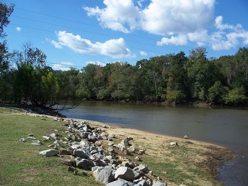 A serene riverbank scene with rocks, sandy shore, and lush trees under a blue sky with fluffy clouds.