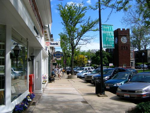 A sunny street scene with shops, parked cars, and a clock tower in the background, showcasing a vibrant small-town atmosphere.