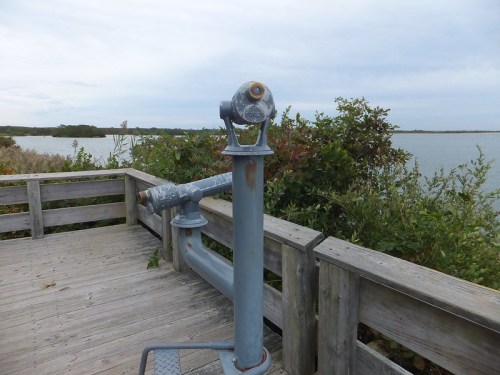A weathered viewing telescope on a wooden deck overlooking a calm body of water and surrounding greenery.