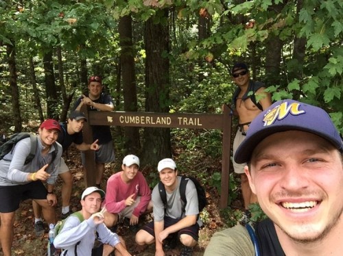 A group of seven hikers poses in front of a Cumberland Trail sign, surrounded by trees and nature.
