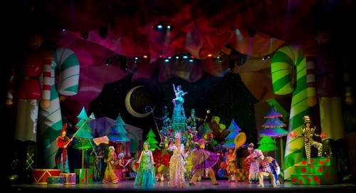 A vibrant stage scene with colorful costumes, festive decorations, and performers in a whimsical holiday-themed show.