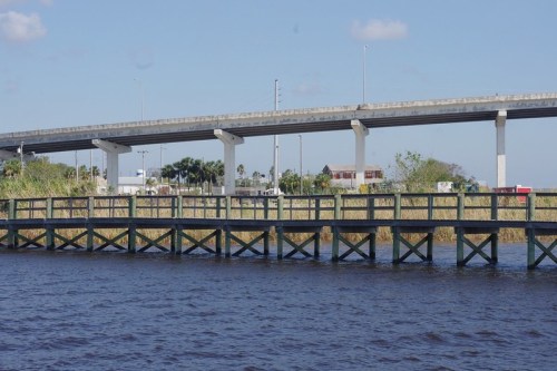 A wooden bridge over water with a highway above, surrounded by greenery and buildings in the background.