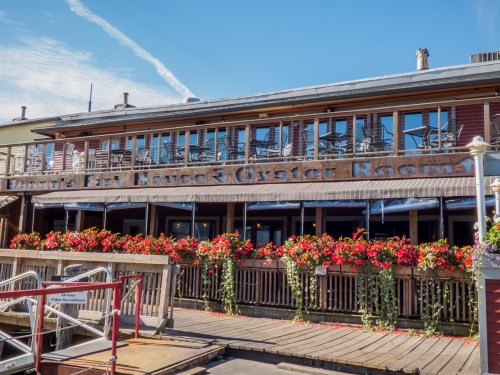 Exterior of a seafood restaurant with a sign, outdoor seating, and vibrant flower boxes under a clear blue sky.