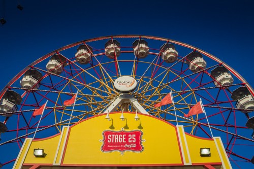 A colorful Ferris wheel against a clear blue sky, featuring a yellow entrance with flags and a "Stage 25" sign.