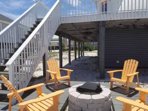 A cozy outdoor seating area with four yellow chairs around a fire pit, under a raised deck on a sandy beach.