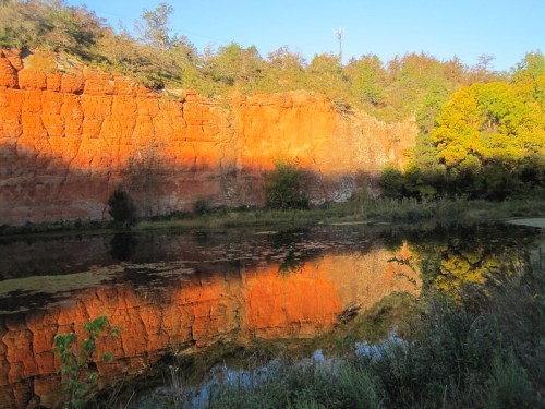 A serene landscape featuring a vibrant orange cliff reflected in a calm pond, surrounded by lush greenery.