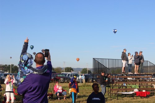A crowd watches hot air balloons in the sky, with one person pointing and taking a photo.