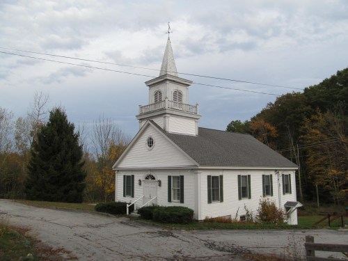 A white church with a steeple sits on a quiet road, surrounded by trees and autumn foliage under a cloudy sky.