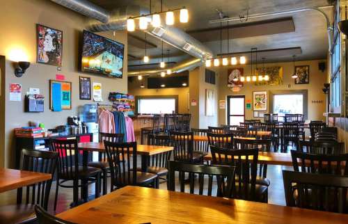 A spacious restaurant interior with wooden tables, black chairs, and a TV showing a basketball game. Colorful artwork on walls.