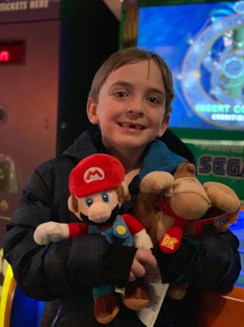 A smiling child holds plush toys of Mario and Donkey Kong in an arcade setting.