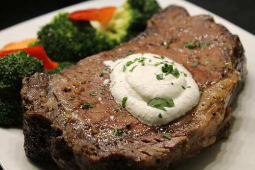 A juicy slice of beef with a dollop of creamy sauce, served with broccoli and red bell peppers on a white plate.