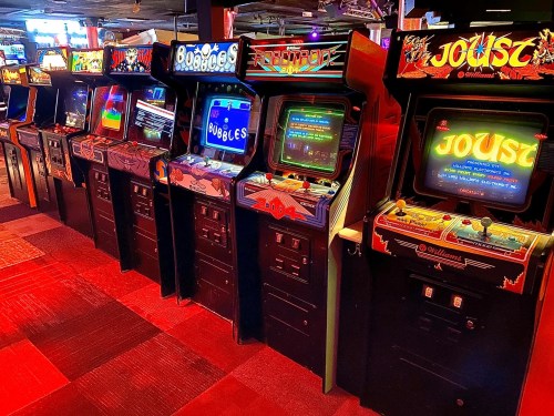 A row of classic arcade game machines, featuring colorful screens and retro designs, in a dimly lit arcade.