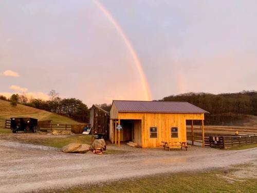 A wooden barn with a metal roof sits on a gravel path, under a rainbow and surrounded by rolling hills.