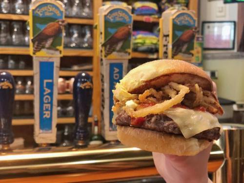A hand holding a burger with melted cheese, crispy onions, and sauce, in front of a bar with beer taps.