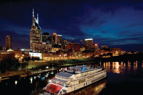 A nighttime view of Nashville's skyline with a riverboat on the water, illuminated buildings, and a dark blue sky.