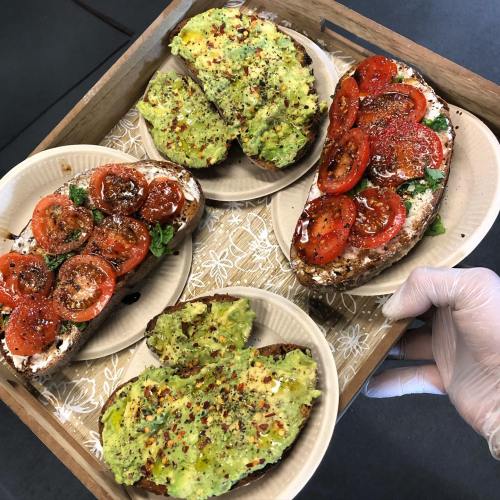 A wooden tray holds four plates of avocado toast topped with tomatoes and spices, served with a gloved hand.