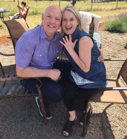 A smiling couple sits together on outdoor chairs, enjoying a sunny day with a bottle of wine in the background.
