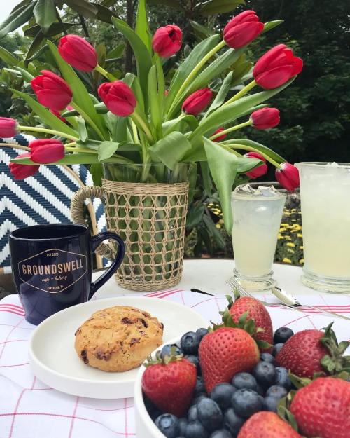 A cozy outdoor table setting with a bouquet of tulips, a coffee mug, a cookie, and bowls of strawberries and blueberries.