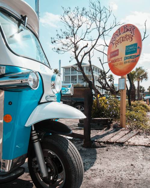 A blue tuk-tuk parked near a sign for Red Bar Returns, surrounded by palm trees and a clear sky.