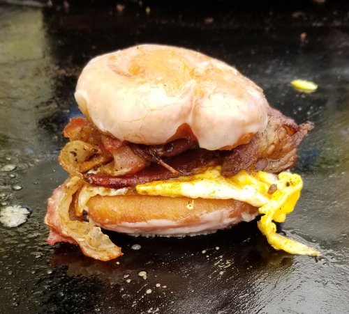 A glazed donut sandwich filled with bacon, sausage, and a fried egg, sizzling on a griddle.